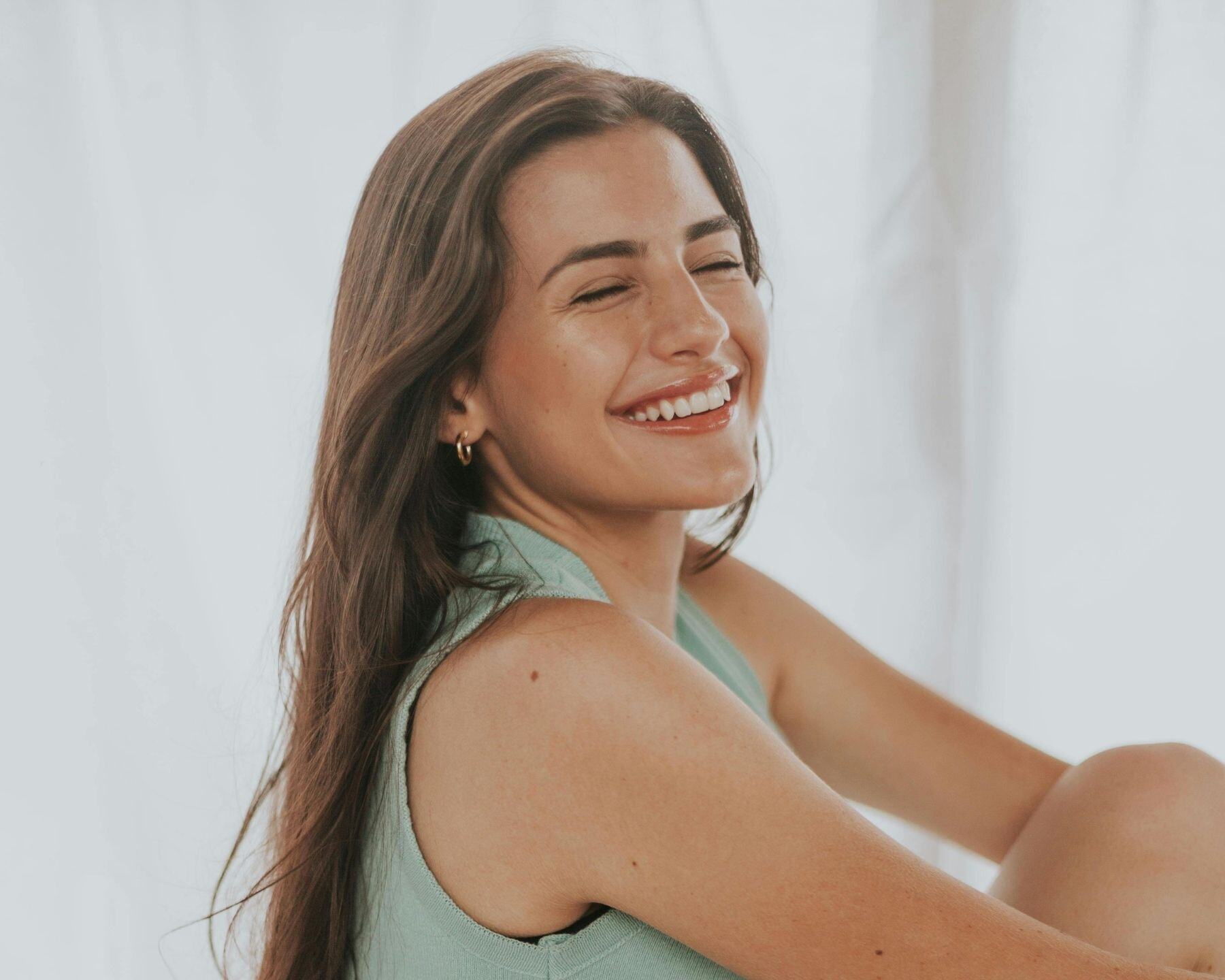 A woman laughing with her eyes closed, against a pale backdrop