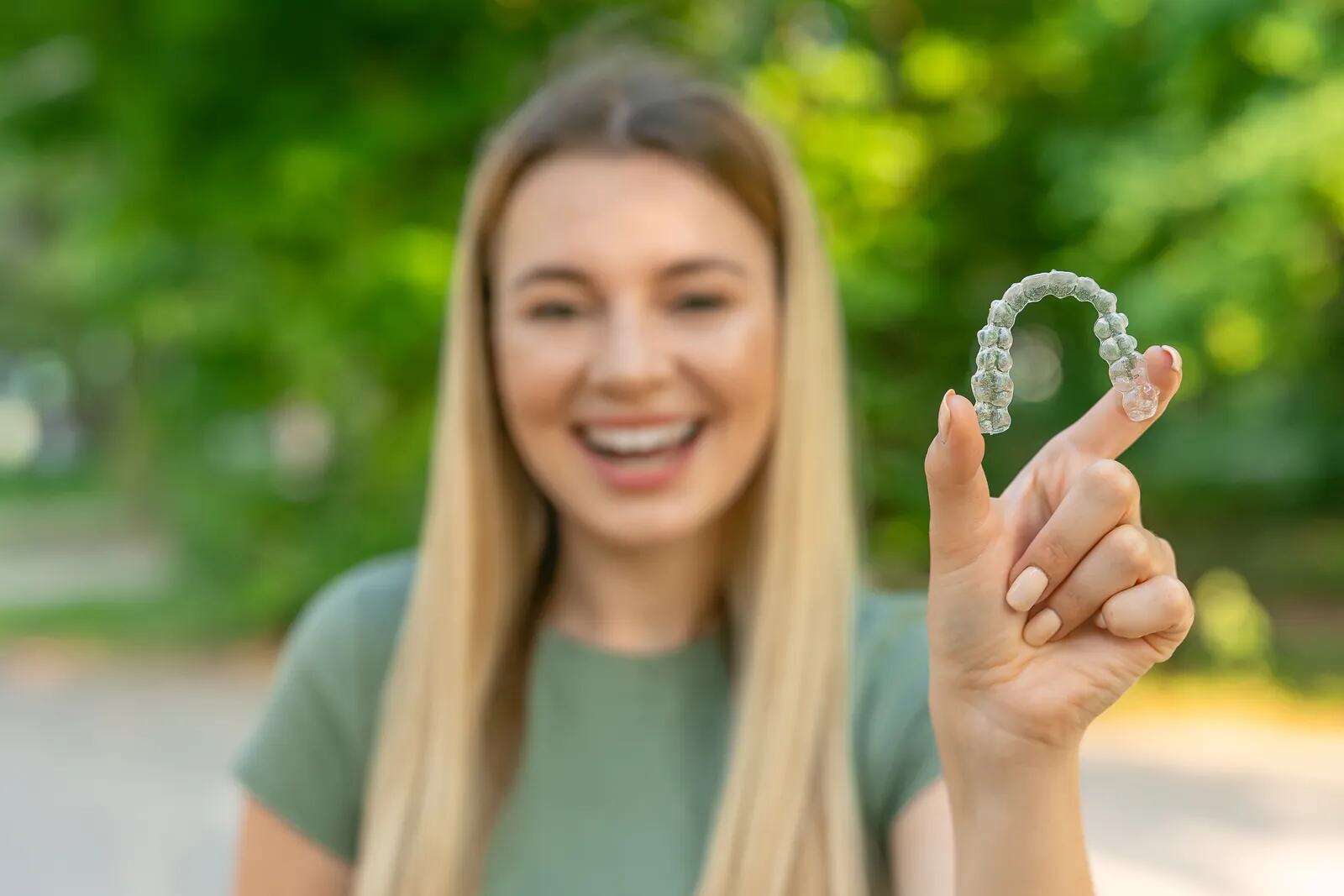 A woman smiling outdoors, holding up her clear aligner